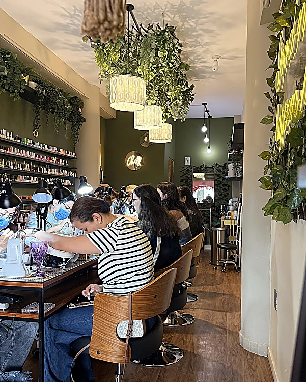 Interior del salón: barra de manicure con sillas de madera, lámparas colgantes con plantas, repisa de esmaltes en la pared y manicuristas trabajando