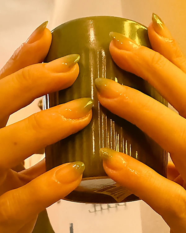 Manos rodeando una taza verde, con uñas almendra de polygel en degradado verde olivo translúcido