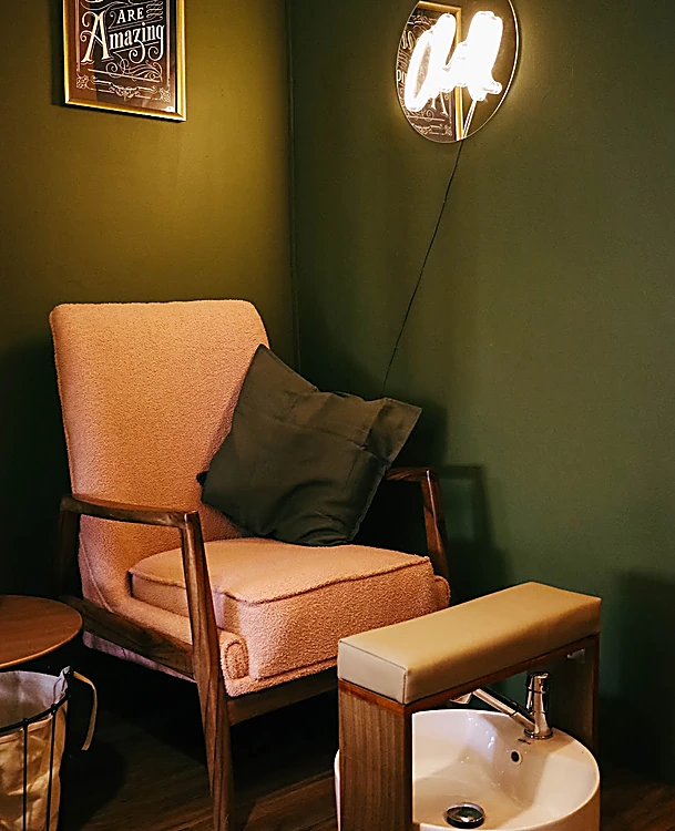 Rincón de pedicure con sillón rosa de madera, cojín verde, lavabo blanco para pies y pared verde oscuro con un cuadro y una lámpara